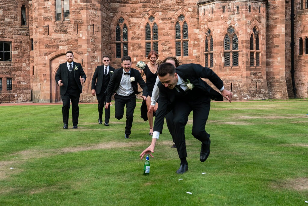 fun groomsmen photo as they all run to grab a bottle of beer on the grass outside Peckforton Castle